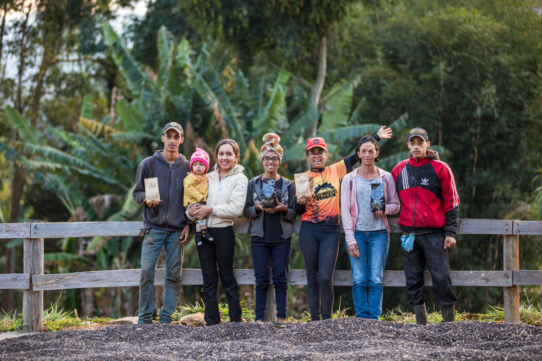 Group of a family with four women, two men, and a toddler standing in front of a fence with drying coffee cherries on the ground.