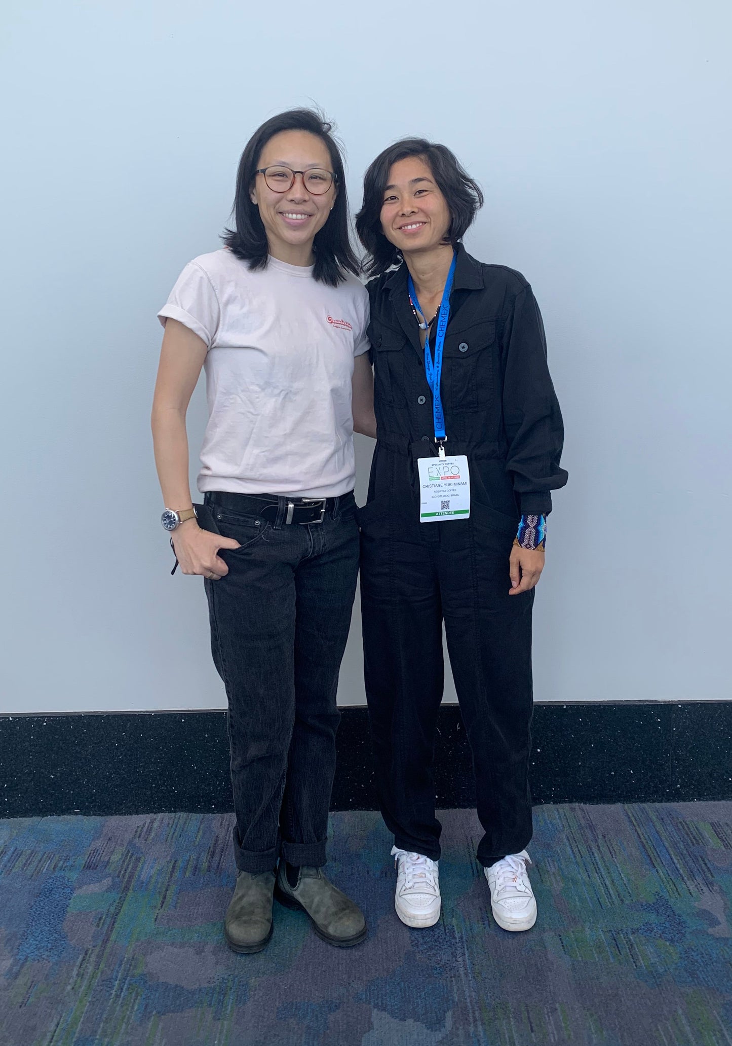 Two women posing and smiling in front of a white wall.