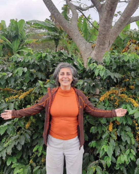 A smiling woman in an orange shirt stands in front of coffee trees with orange coffee cherries on its branches.
