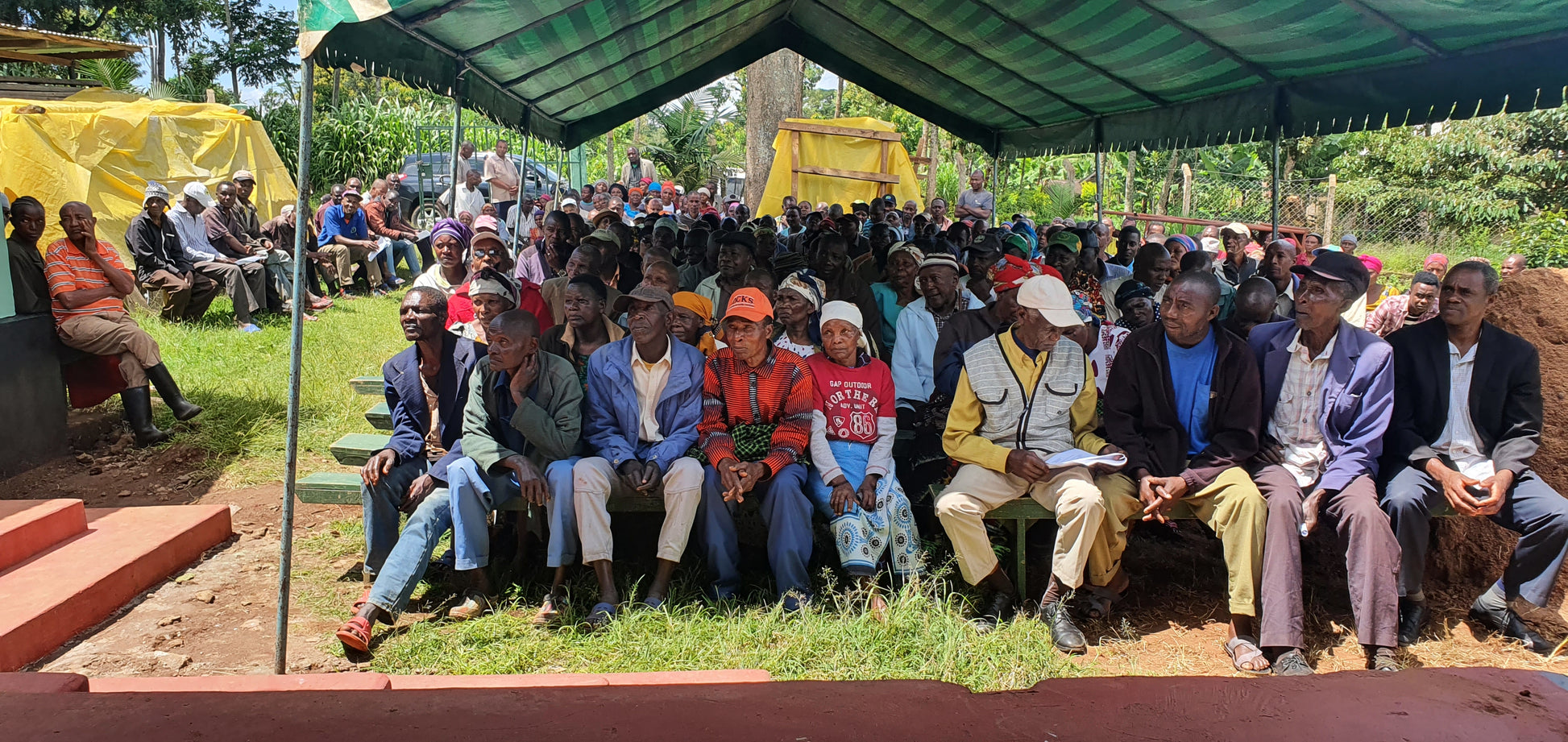 Community members sit, gathered under a large canopy tent.